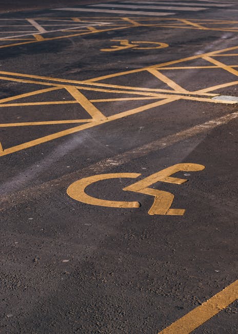 A close-up view of a parking lot surface showing yellow painted markings, including accessible parking symbols and designated spaces outlined with diagonal lines. The asphalt appears dark and slightly textured, with some white markings possibly indicating additional parking instructions. The visible area suggests an outdoor environment typical of a residential or commercial building vicinity, where relocation services by Man With a Van Peckham might operate. The markings are well-maintained, clearly visible, and designed to regulate vehicle parking during home removals or furniture transport. This scene exemplifies the logistical considerations involved in loading and unloading within designated parking zones for house relocation, with the space suitable for a removal van accessed during the ongoing or planned moving process as described on the 'Bellenden Road Removals' page.