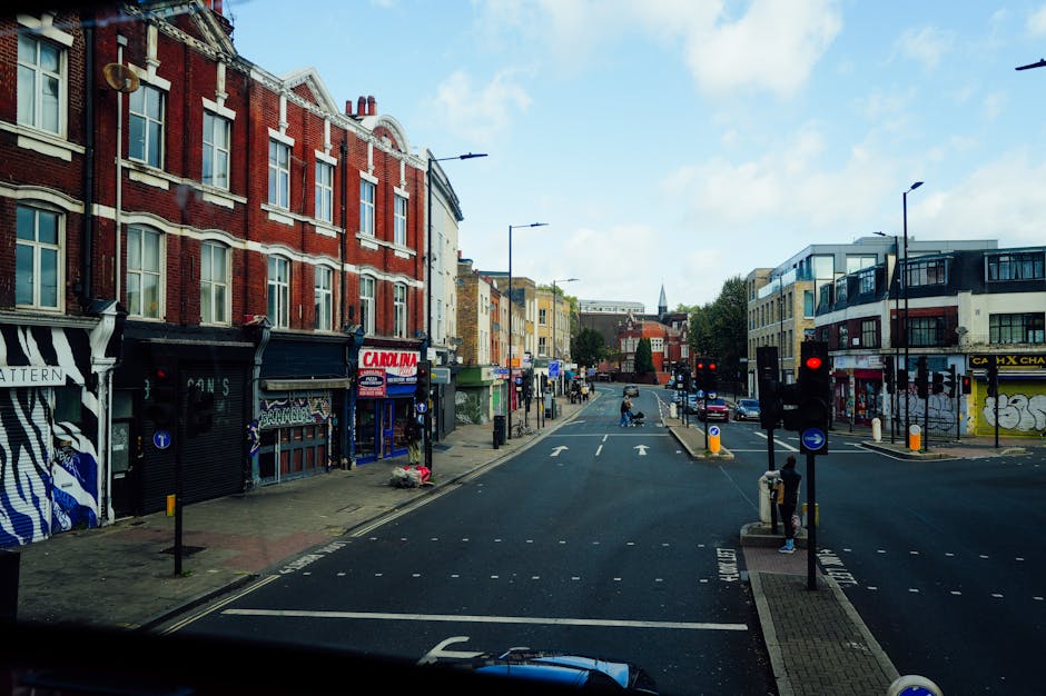 A daytime street scene on Bellenden Road in Peckham showing a row of buildings with retail shops on the ground floor, some with painted facades and signage, and residential windows above. The street has traffic lights, road markings, and designated parking zones. The pavement is lined with bollards, and pedestrians are waiting at the corner. On the left side, storefronts include a shop with black and white striped artwork and another with a 'Carolina' sign. The scene captures an active area suitable for home relocation and furniture transport activities, with the street providing space for loading and unloading during removals, as managed by Man With a Van Peckham.