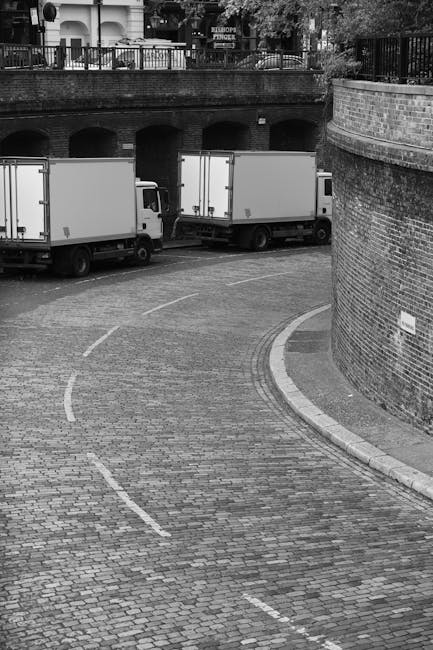 A black-and-white photograph shows a curved cobblestone street in an urban area with two white moving vans parked next to each other on the road, partially underneath an arched brick bridge. The vans