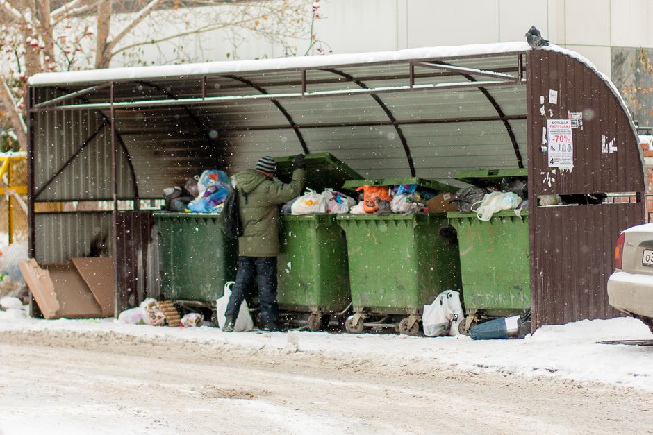 An outdoor scene of overflowing waste and rubbish bags piled around a large mixed paper and cardboard recycling container, situated on a paved pavement in front of a commercial building. The waste inc