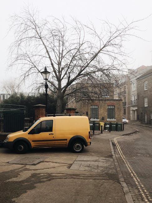 A white commercial van parked on a city street in front of a multi-storey building with beige or light grey facades and narrow windows. The van's rear doors are open, revealing a cluttered load of lar