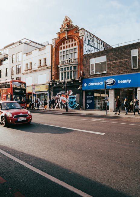 A wide view of Queens Road in Peckham during daytime, featuring a row of multi-storey terraced houses with brick facades and pitched roofs, some with bay windows. The street is busy with several cars