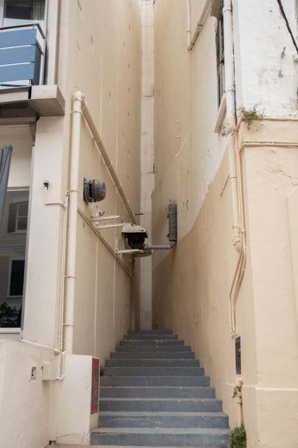A narrow outdoor staircase with grey steps ascending between two tall beige building walls, which show visible pipes, utility boxes, and electrical meters attached to the surfaces. The staircase is situated in an alleyway or back passage, with minimal natural light illuminating the scene. The walls are bare except for small patches of plant growth near the right side, and there is no visible handrail. This setting reflects a typical access point for a home or apartment building used during a house relocation or furniture transport process, consistent with packing and moving activities in a dense urban environment. The image potentially captures a scene where removal specialists like Man With a Van Peckham may need to navigate narrow stairs during the loading or unloading stages of a house removal. The overall scene emphasizes logistical challenges faced during furniture transport through confined access points on a residential property in SE15, Peckham.