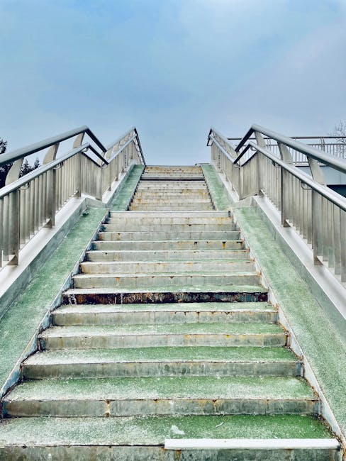 A narrow outdoor staircase with grey steps ascending between two tall beige building walls, which show visible pipes, utility boxes, and electrical meters attached to the surfaces. The staircase is situated in an alleyway or back passage, with minimal natural light illuminating the scene. The walls are bare except for small patches of plant growth near the right side, and there is no visible handrail. This setting reflects a typical access point for a home or apartment building used during a house relocation or furniture transport process, consistent with packing and moving activities in a dense urban environment. The image potentially captures a scene where removal specialists like Man With a Van Peckham may need to navigate narrow stairs during the loading or unloading stages of a house removal. The overall scene emphasizes logistical challenges faced during furniture transport through confined access points on a residential property in SE15, Peckham.