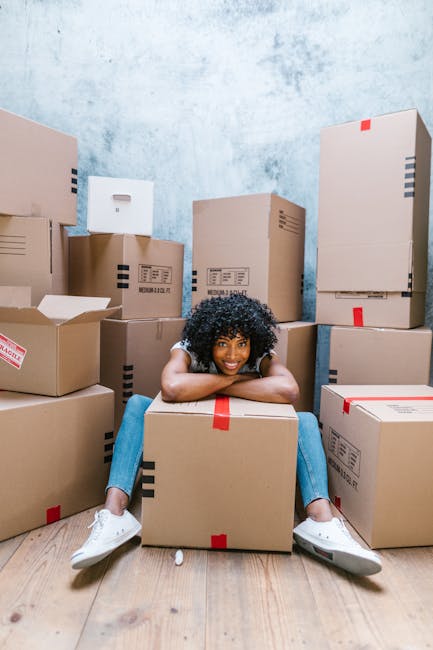 A young woman with curly black hair, dressed in casual clothing and white sneakers, is sitting on the floor inside a room surrounded by stacked cardboard moving boxes, some sealed with red or black ta