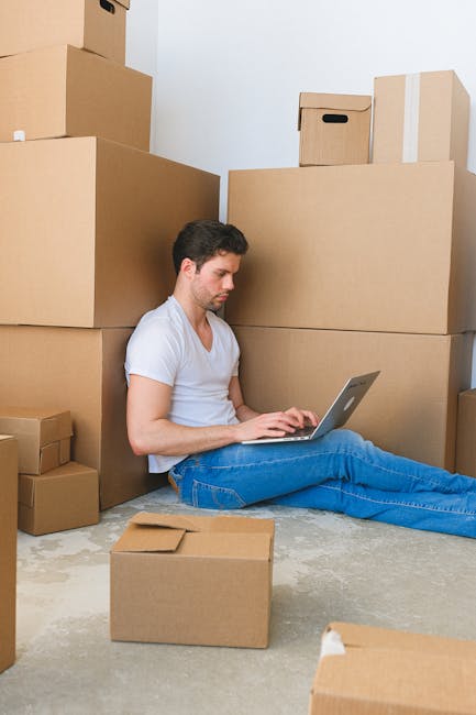 A young woman with curly black hair, dressed in casual clothing and white sneakers, is sitting on the floor inside a room surrounded by stacked cardboard moving boxes, some sealed with red or black ta